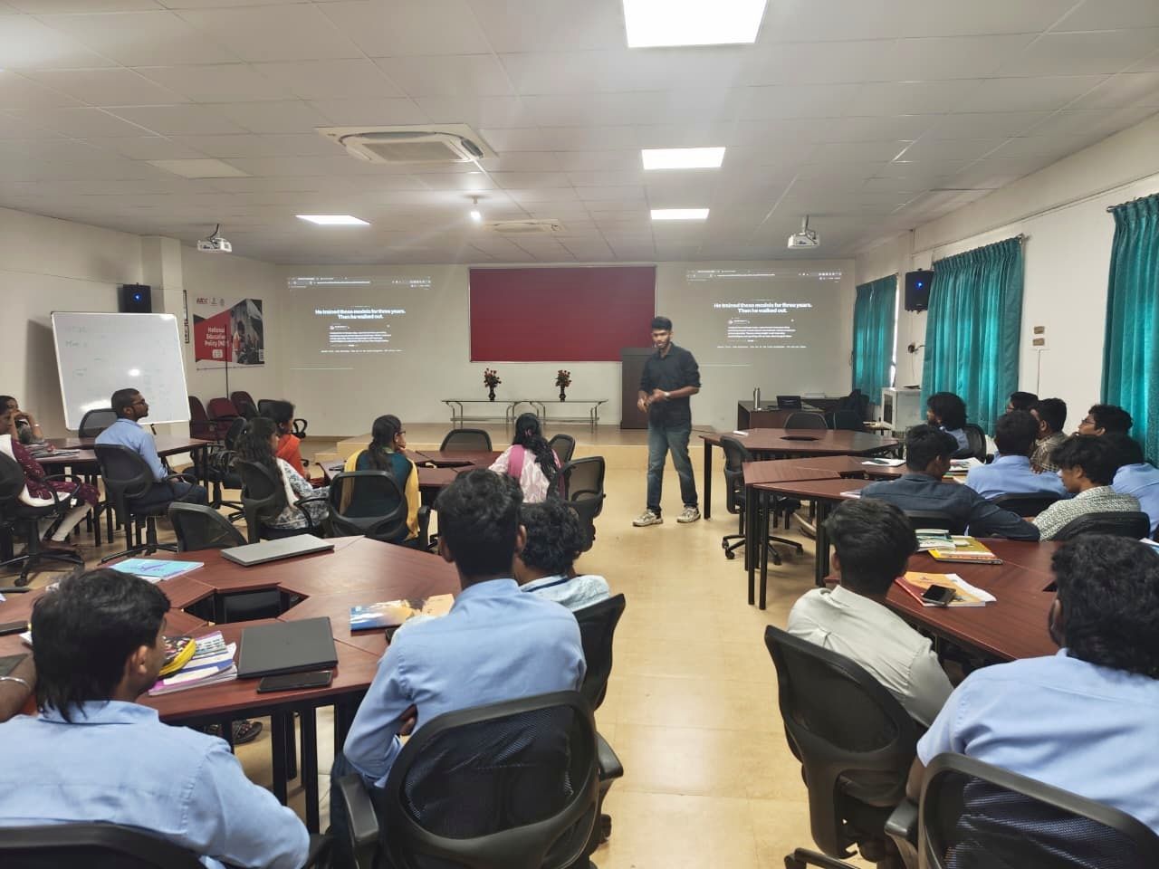 Kanish speaking at the front of the classroom, students seated around tables