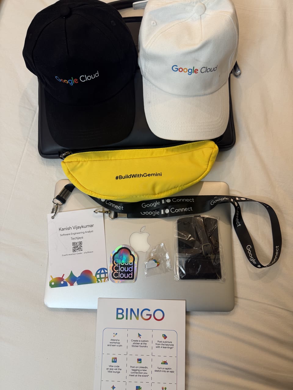 Two Google Cloud caps, a yellow #BuildWithGemini sling bag, an I/O Connect lanyard and badge, stickers and a BINGO card